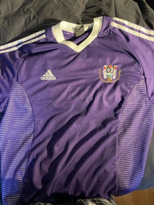 Vintage Anderlecht Fotbollströja L - Säljer en lila fotbollströja från Adidas med vita detaljer och klubbmärke på bröstet. Tröjan har vita ränder på axlarna, rund halsringning med vit kant och meshpaneler på sidorna för extra ventilation. Perfekt för träning eller match.