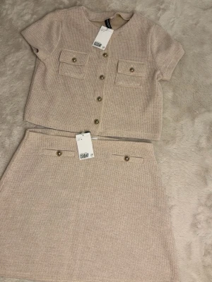 Beige stickat set med kjol och topp - Snyggt tvådelat set från H&M i beige stickat material. Toppen har korta ärmar, dekorativa bröstfickor och guldknappar framtill. Kjolen är rak och har dekorativa fickor med matchande knappar. Perfekt för en trendig och stilren look.