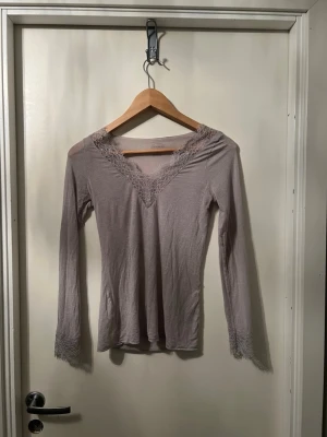Intimissimi tröja - Säljer en ljuslila/grå långärmad topp med v-ringning och fina spetsdetaljer vid halsringningen och ärmsluten. Toppen har en slim passform och är tillverkad i ett mjukt material som känns skönt mot huden. Perfekt för dig som gillar romantiska detaljer. Aldrig använd. Pris kan diskuteras