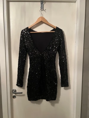 Svart paljettklänning med öppen rygg  - Snygg svart kortklänning täckt i glittriga paljetter. Klänningen har lång ärm och djup v-ringning bak, vilket ger en festlig look. Figurnära passform och stretchigt material som formar sig efter kroppen. Perfekt för dig som vill sticka ut på festen!