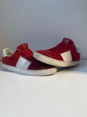 Valentino Flycrews  - Snygga röda sneakers från Valentino med vita detaljer och röd mocka. Skorna har vita partier på sidan, vita sulor, röda skosnören och nitar där bak vid hälen. Yttermaterialet är en mix av mocka och läder, och sulan har ett unikt mönster undertill. Perfekta för dig som vill sticka ut.