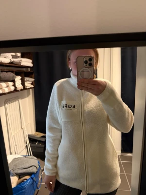 Vit teddyjacka från EQPE, M - Mysig vit teddyjacka från EQPE med dragkedja framtill och hög krage. Jackan har en bröstficka med EQPE-logga och är gjord i fluffigt teddy-material som håller dig varm. Perfekt för lager på lager och chill stil.