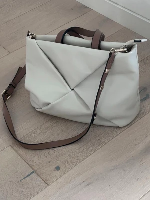 Handväska från Parfois - Härligt följsam väska i ljust konstläder med bruna detaljer. Storlek 35x24. Väskan är rymlig och har ett innerfack med dragkedja. Den är i bra skick men lite sliten på handtagen. Se sista bilden.