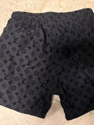 Mörkblå Louis Vuitton badbyxor - Säljer ett par mörkblå badbyxor från Louis Vuitton med klassiskt monogrammönster i svart. De har elastisk midja och är tillverkade i ett lätt, snabbtorkande material. Perfekta för stranden eller poolen.