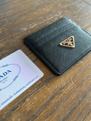 Svart kortfodral - Stilrent svart kortfodral  i strukturerat l med guldfärgad logga framtill. Fodralet har flera kortfack och en smidig, tunn design som passar perfekt i fickan. Klassisk och lyxig accessoar för dig som gillar exklusiva detaljer. Helt ny 