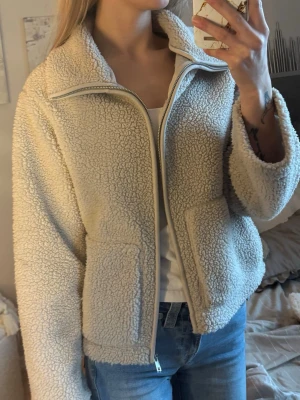 Beige teddyjacka med dragkedja - Mysig beige teddyjacka med hög krage och dragkedja framtill. Jackan har två stora fickor och är gjord i fluffigt teddy-material som ger en avslappnad och trendig look. Perfekt att slänga på sig över en hoodie eller t-shirt för extra värme.