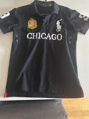 Svart Ralph Lauren Chicago piké - Snygg svart pikétröja från Ralph Lauren med stora broderade detaljer, CHICAGO-text framtill och nummer 3 på ryggen. Klassisk krage, korta ärmar och vita kontrastdetaljer. Perfekt för dig som gillar sportig och stilren look. Chief keef polo