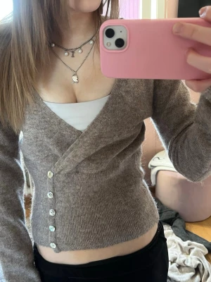 Beige omlottkofta med knappar - Snygg beige omlottkofta med v-ringning och dekorativa knappar längs sidan. Koftan har lång ärm och är stickad i ett mjukt material som ger en avslappnad men trendig look. Perfekt att styla med ett linne under för en lager-på-lager känsla.