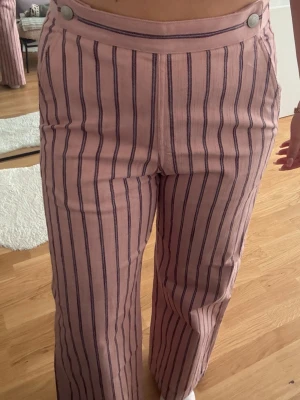 Rosa randiga linnebyxor & Other Stories - Säljer ett par rosa linnebyxor från & Other Stories med mörkblå vertikala ränder. Byxorna har hög midja, vida ben och två silverfärgade knappar i linningen. Materialet känns luftigt och perfekt för varma dagar. Snyggt mönster som sticker ut.