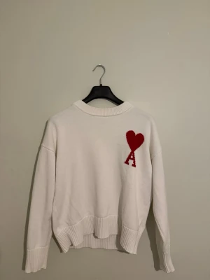 Ami De Paris - Vit stickad tröja från Ami Paris med röd hjärtlogga och bokstaven A på bröstet. Tröjan har rund hals, ribbade muddar och avslut samt en avslappnad passform. Perfekt för dig som gillar stilrena plagg med en twist.