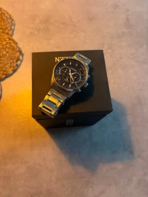 Citizen herrklocka i silver - Stilren herrklocka från Citizen med silverfärgad länk i metall och svart urtavla med flera funktioner. Klockan har klassisk rund form och levereras med extra länkbitar för justering av storlek. Perfekt accessoar för en modern look.