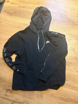 Svart Nike hoodie med logga - Säljer en svart hoodie från Nike med vit logga på bröstet och band med Nike-tryck längs ärmarna. Hoodien har en stor huva (glansig) med dragsko och en klassisk magficka. 