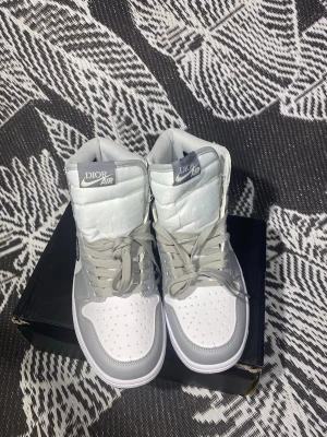 Dior x Air Jordan 1 High sneakers - Exklusiva Dior x Air Jordan 1 High sneakers i vitt och ljusgrått läder med grå skosnören och tydlig Dior Air-logga på plösen. Begränsad upplaga med numrering på insidan. Snygg och ikonisk siluett som sticker ut.