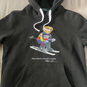 Svart hoodie Polo Bear Ralph Lauren - Svart hoodie från Ralph Lauren med Polo Bear-tryck framtill där björnen åker skidor. Klassisk huva med vita snören och känguruficka. Mjuk bomullskvalitet och avslappnad passform. Perfekt för dig som gillar streetwear och ikoniska prints. Storlek L men passar även M. Pris kan diskuteras vid snabb affär