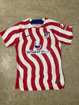 Atletico Madrid matchtröja Nike - Säljer en Atletico Madrid matchtröja från Nike med röda och vita vågiga ränder, klubbmärke på bröstet och WhaleFin sponsortryck. Tröjan har korta ärmar och är tillverkad i ett lätt, ventilerande material som passar perfekt till fotbollsträning eller match.