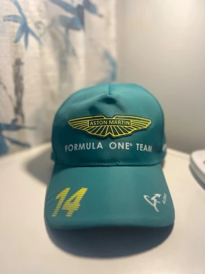 Aston Martin Formula One keps - Fernando Alonso - Snygg grön keps från Aston Martin Formula One Team med Alonso-tryck och nummer 14 på skärmen. Kepsen har broderad logga framtill och coola vita och gula detaljer. Perfekt för dig som gillar racing och vill sticka ut på läktaren.