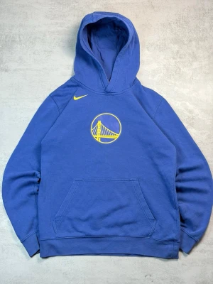 Blå Golden State Warriors hoodie Nike - Snygg blå hoodie från Nike med Golden State Warriors-logga i gult på bröstet och gul Nike swoosh på axeln. Klassisk känguruficka framtill och stor huva. Tillverkad i mjukt bomullsmaterial, perfekt för dig som gillar basket och streetwear.