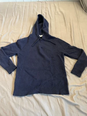 Mörkblå stickad hoodie från Oscar Jacobson - Mörkblå stickad hoodie från Oscar Jacobson med huva och dragsko. Tillverkad i mjukt material som ger en avslappnad och stilren look. Långa ärmar och ribbade muddar vid ärmslut och nederkant. Perfekt för dig som gillar en enkel men snygg stil.