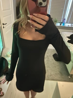 Svart långärmad kortklänning - En stilren svart kortklänning med lång ärm och fyrkantig urringning. Klänningen har en tight passform och är perfekt för dig som gillar minimalistisk och trendig stil. Materialet känns mjukt och följsamt mot kroppen.