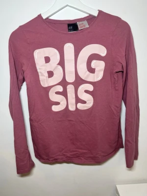 Rosa långärmad pyjamaströja H&M - Mysig långärmad pyjamaströja från H&M i dammigt rosa med stort tryck 'BIG SIS' i ljusrosa framtill. Tillverkad i mjuk bomull, perfekt för avslappnade kvällar hemma. Rund halsringning och bekväm passform.