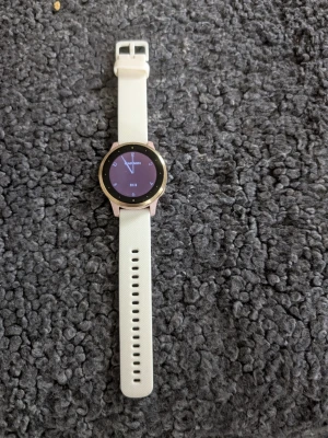 Garmin smartklocka med vit silikonrem - Snygg smartklocka från Garmin med rund urtavla och vit silikonrem. Klockan har guldfärgad ram och rosa detaljer runt boetten. Pekskärm med digital visning och klassisk design. Perfekt för dig som vill ha koll på både tid och notiser direkt på handleden.