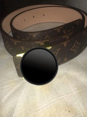 Bälte - Snyggt brunt bälte med klassiskt monogrammönster och guldfärgat spänne. Bältet har flera hål för justerbar passform och insidan är ljusbeige. Perfekt accessoar för att lyfta din outfit.