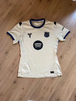 FC Barcelona bortatröja beige player edition - Snygg beige FC Barcelona bortatröja från säsong 25/26 med svarta och blå detaljer. Tröjan har klubbmärke, Spotify-logga och UNHCR-tryck. Tillverkad i lätt och ventilerande material, perfekt för fotboll eller träning. Rund halsringning och korta ärmar. Storlek L. Pris kan diskuteras vid snabb affär.