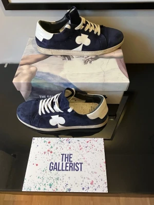 Blå-vita sneakers från The Gallerist - Snygga sneakers från The Gallerist i mörkblå mocka med vita detaljer och vita skosnören. Skorna har en unik klöverdetalj på sidan och vit hälkappa. Perfekta för dig som vill sticka ut med stilrena skor. Box ingår.