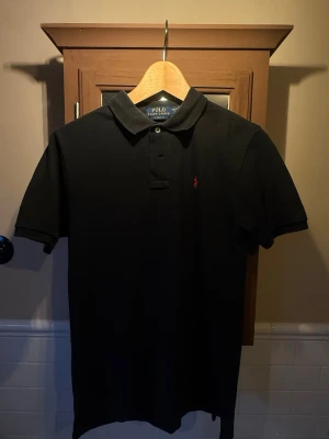 Svart pikétröja från Polo Ralph Lauren - Klassisk svart pikétröja från Polo Ralph Lauren med korta ärmar och krage. Tröjan har knappar vid halsen och den ikoniska röda logotypen broderad på bröstet. Perfekt till jeans eller shorts för en clean och stilren look. Modellen är i storlek barn 14-16 år, fast borde passa i storlek xs. T-shirten är i fint skick men har lite slitage i kragen som, man knappast märker (kan se på bild 3). 