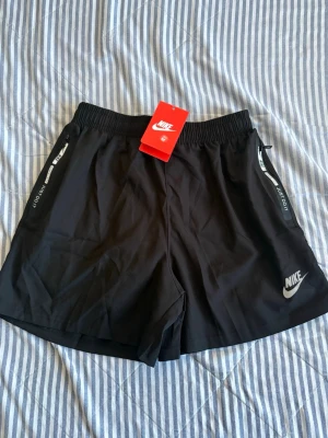 Svarta träningsshorts från Nike - Snygga svarta korta träningsshorts från Nike med vitt logotryck på benet och textdetaljer längs sidorna. Shortsen har elastisk midja och är tillverkade i ett lätt och snabbtorkande material, perfekta för aktiva dagar.