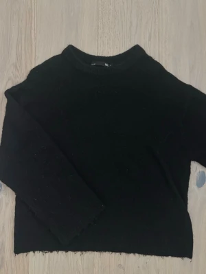 Svart stickad tröja från 157 - En enkel och stilren svart stickad tröja från 157 med rund halsringning och långa ärmar. Perfekt basplagg till garderoben och lätt att matcha med olika outfits. Tröjan har en mjuk känsla och ribbade muddar vid halsen.