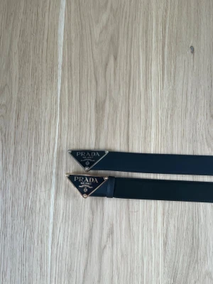 Svart bälte från hm  med metallspänne - Snyggt svart bälte från hm med ikoniskt trekantigt metallspänne i silver och guld. Bältet har en stilren design och är tillverkat i slätt skinn. Perfekt accessoar för att lyfta vilken outfit som helst.