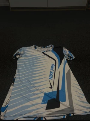 Nike Pro blåvit t-shirt med byxor  - Nike Pro t-shirt i blått, vitt och grått med grafiskt mönster och stor logga framtill. Kortärmad modell i lätt och stretchigt material, perfekt för träning eller sportiga outfits. Snygg passform och modern design.