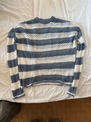 Randig stickad tröja från Bershka - Säljer en stickad tröja från Bershka i storlek S. Tröjan har breda horisontella ränder i ljusblått och vitt samt ett hålmönster över hela plagget. Långärmad modell med ribbstickad hals, ärmslut och nederkant. Perfekt för lager på lager och en avslappnad stil.