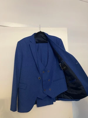 Blå kostym med väst, kavaj och byxor strl 48 - Snygg blå kostym med matchande väst, kavaj och byxor. Kostymen har ett diskret mönster, dubbla knapprader på västen och klassisk passform. Använd vid ett tillfälle