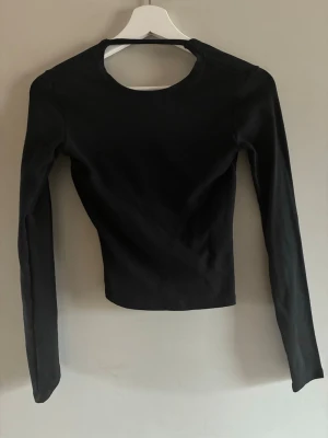 Svart långärmad omlottopp - Trendig svart långärmad topp med omlottdetalj framtill och rundad halsringning. Toppen har en tight passform och är tillverkad i ett stretchigt material som känns bekvämt mot huden. Perfekt för dig som gillar minimalistisk och stilren design.