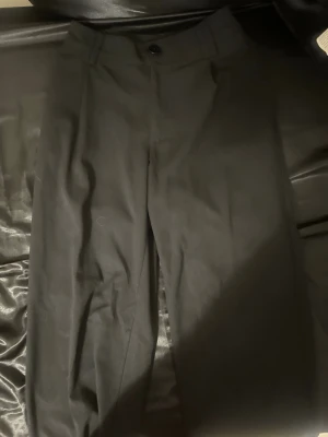 Svarta kostymbyxor med hög midja - Snygga svarta kostymbyxor med hög midja och raka ben. Byxorna har en klassisk knappstängning framtill och bälteshällor. Perfekta för en stilren och clean look. Materialet ser ut att vara en mjuk och lätt vävd textil som ger en dressad känsla.