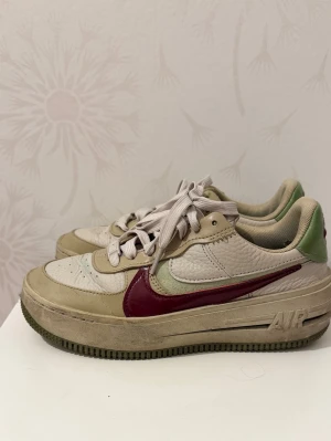 Nike Air Force 1 beige/grön/vinröd - Nike Air Force 1 sneakers i beige med detaljer i grönt och vinrött. Skorna har en klassisk låg siluett, vit mesh på tån och sidorna samt ett vinrött swoosh-märke. Yttermaterialet är en mix av läder och textil. Grön detalj på hälen och beige snörning.