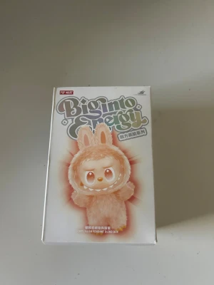 Pop Mart Big Into Energy figur - Säljer en Pop Mart Big Into Energy blind box med en söt figur i beige plyschdräkt med kaninöron. Förpackningen är dekorerad med färgglad text och illustration av figuren. Perfekt för samlare av designerleksaker eller som present. Nu får du chansen att öppna paketet och få en random figur från Labubu!