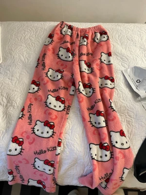 Hello Kitty rosa fleecebyxor - Mysiga rosa loungewear-byxor i fleece med Hello Kitty-motiv och text. Byxorna har resår i midjan och är täckta av vita Hello Kitty-ansikten med röda rosetter. Perfekta för chill hemma eller sleepovers.