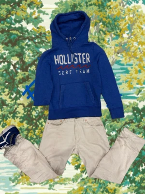Blå Hollister hoodie med tryck - Blå hoodie från Hollister med vit och röd text 'Hollister Surf Team' på bröstet. Klassisk känguruficka framtill, dragsko i huvan och mjukt material på insidan. Perfekt för en avslappnad och sportig stil.