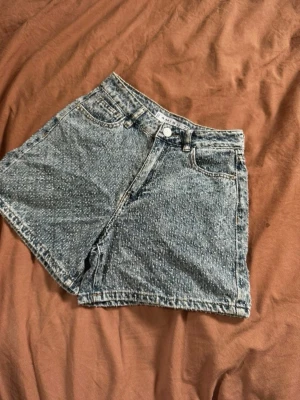 Högmidjade blå jeansshorts - Säljer ett par klassiska högmidjade jeansshorts i blå denim. Shortsen har fem fickor, bälteshällor och stängs med knapp och dragkedja. Perfekta till sommaren och ger en avslappnad vintagekänsla.