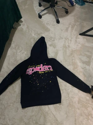 Svart hoodie med spindelnät och stjärnor - Cool svart hoodie med stor huva och tryck framtill. Texten 'spider' i rosa och vitt, omgiven av gula och lila stjärnor samt ett stort spindelnät. Perfekt för dig som gillar streetwear och unika prints. Materialet känns mjukt och bekvämt, och huvan är rymlig.