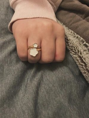 Guld ring med vita detaljer - Snygg guld ring med två vita detaljer, en i form av en blomma och en sexkantig. Ringen har en modern och trendig design som sticker ut och passar perfekt för dig som gillar unika accessoarer. Materialet ser ut att vara metall med emaljliknande yta.