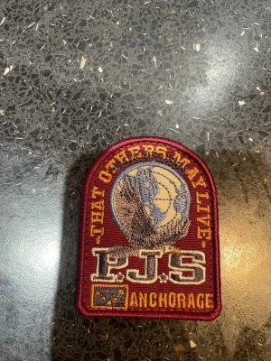 Patch P.J.S Anchorage brodyrmärke - Snyggt broderat patch med texten 'THAT OTHERS MAY LIVE' och 'P.J.S ANCHORAGE'. Märket har en rundad form, vinröd bakgrund och detaljer i gult, grått och vitt. Perfekt att fästa på jacka eller väska för en cool look. Storleken är 6,5x5cm. Passar till vinterjacka modeller för kvinnor med pälskrage.