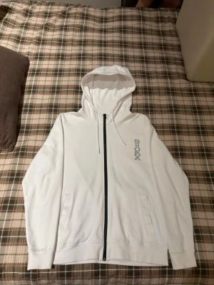 Vit Hugo zip hoodie med tryck  - Säljer en clean vit hoodie med dragkedja och huva. På bröstet och ryggen finns ett stilrent svart tryck med grafiskt mönster. Hoodien har fickor på sidorna och ribbade muddar vid ärmslut och nederkant. Perfekt för dig som gillar minimalistisk streetwear.