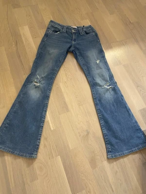 Blå utsvängda jeans med slitningar från Gina - Säljer ett par lågmidjade, blå, utsvängda jeans med slitningar på knäna och snygga glittriga broderade detaljer på bakfickorna. Jeansen är i storlek 164 och knappt använda.