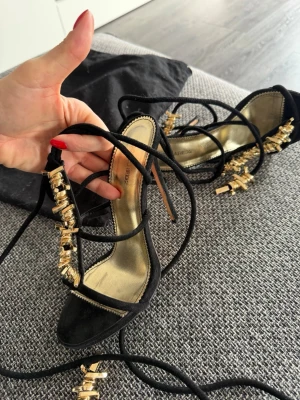 Svarta sandaler med gulddetaljer Dsquared2 - Snygga svarta sandaler från Dsquared2 med tunna remmar och dekorativa guldfärgade detaljer. Sandalerna har öppen tå och snörning som lindas runt benet. Innersulan är i guldton och skon har en elegant, feminin design. Perfekta för att lyfta vilken outfit som helst.