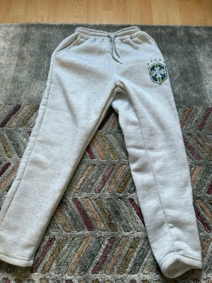 Grå mjukisbyxor med Saint-logga - Snygga grå mjukisbyxor från Saint med broderad logga och fem gröna stjärnor på vänster lår. Byxorna har resår i midjan med dragsko och två sidofickor. Perfekta för chill dagar eller när du vill ha det bekvämt.