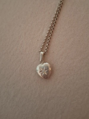 Silverhalsband med hjärthänge - Snyggt halsband i silverfärg med ett litet hjärthänge. Kedjan är tunn och klassisk, och hjärtat har en diskret stjärndetalj på framsidan. Perfekt för dig som gillar stilrena och enkla smycken.